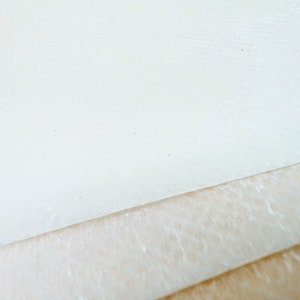 May include: Close-up of three layers of white fabric. The top layer is smooth, the middle layer has a textured surface, and the bottom layer is a smooth, light brown fabric.