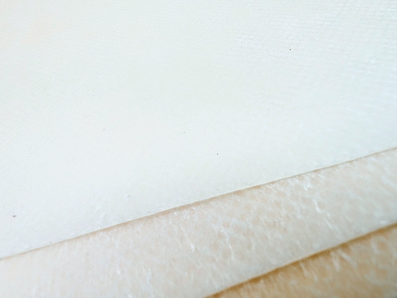 May include: Close-up of three layers of white fabric. The top layer is smooth, the middle layer has a textured surface, and the bottom layer is a smooth, light brown fabric.