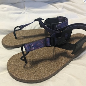 Puede incluir: Un par de sandalias de corcho con correas ajustables. Las correas son negras con una tela estampada en morado y rosa.