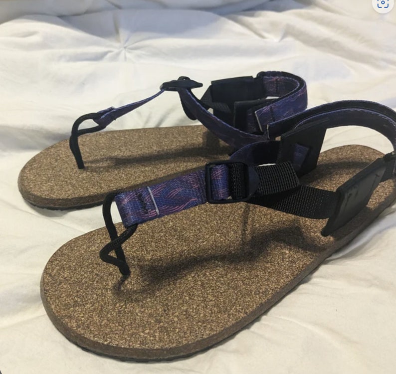 Puede incluir: Un par de sandalias de corcho con correas ajustables. Las correas son negras con una tela estampada en morado y rosa.