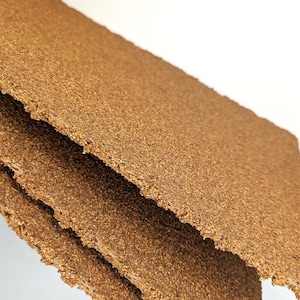 May include: Close-up of a stack of brown, textured, rectangular cork tiles. The tiles are made of natural cork and have a rough, uneven surface.