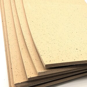 May include: A stack of rectangular cork sheets. The sheets are a light tan colour with small, dark speckles throughout. The edges of the sheets are a darker brown, and the sheets are stacked on top of each other.