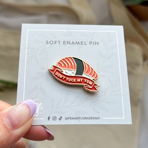 May include: A gold-coloured enamel pin with a red and black sushi design. The pin has a white banner with the text "Don't Yuck My Yum".