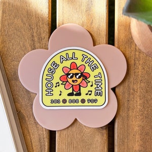 May include: A pink flower-shaped coaster with a yellow sticker featuring a cartoon flower wearing sunglasses and the text "House All The Time 303 808 909".
