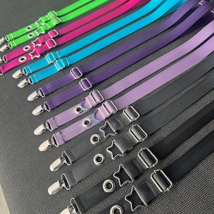 May include: A collection of adjustable elastic straps in neon green, hot pink, bright blue, purple, lavender, and black. Each strap features a silver metal clip at one end, a star-shaped metal slider, and a circular metal eyelet for length adjustment.