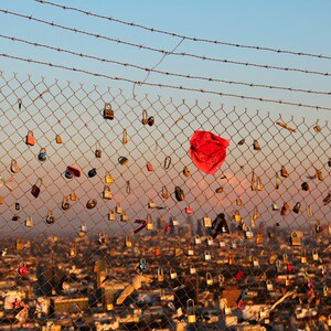 May include: A chain link fence with barbed wire at the top is covered in padlocks. A red bandana hangs from the fence. The fence is in front of a city skyline.