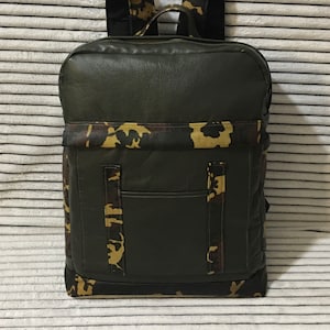 May include: A dark green backpack with camouflage detailing on the straps, front pocket, and base. The backpack is rectangular with a zip closure and a front pocket. The straps are padded for comfort.