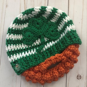 May include: A green and white crocheted hat with a shamrock design and an orange crocheted beard. The hat has a button on the side.