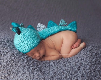 May include: A newborn baby wearing a blue and green crocheted dinosaur costume, sleeping on a gray fuzzy blanket.