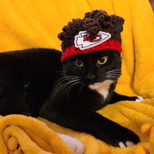 May include: A black and white cat wearing a brown and red crocheted hat with the Kansas City Chiefs logo. The cat is lying on a yellow blanket with pom-poms.