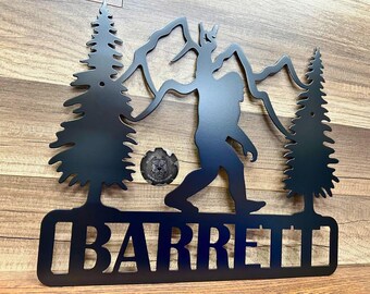 May include: Black metal sign featuring a Bigfoot silhouette walking across a landscape with mountains and trees. The name "BARRETT" is displayed in bold letters at the bottom. A circular emblem is also present.