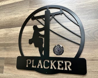 May include: Black metal wall art featuring a lineman climbing a utility pole, set within a circular frame. The word "PLACKER" is cut out at the bottom. A small emblem is visible below the frame.
