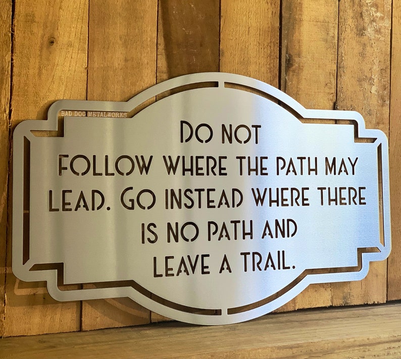 Puede incluir: Un letrero de metal plateado con el texto "NO SIGAS EL CAMINO. VE EN CAMBIO DONDE NO HAY CAMINO Y DEJA UN RASTRO". El letrero tiene una forma decorativa y est&aacute; montado sobre una superficie de madera.