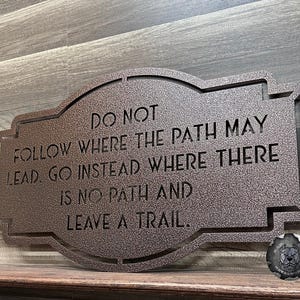 Puede incluir: Un letrero de metal de color bronce con la cita: "NO SIGAS EL CAMINO. VE EN CAMBIO DONDE NO HAY CAMINO Y DEJA UN RASTRO." El letrero tiene un borde decorativo y es adecuado para exhibici&oacute;n en interiores o exteriores.