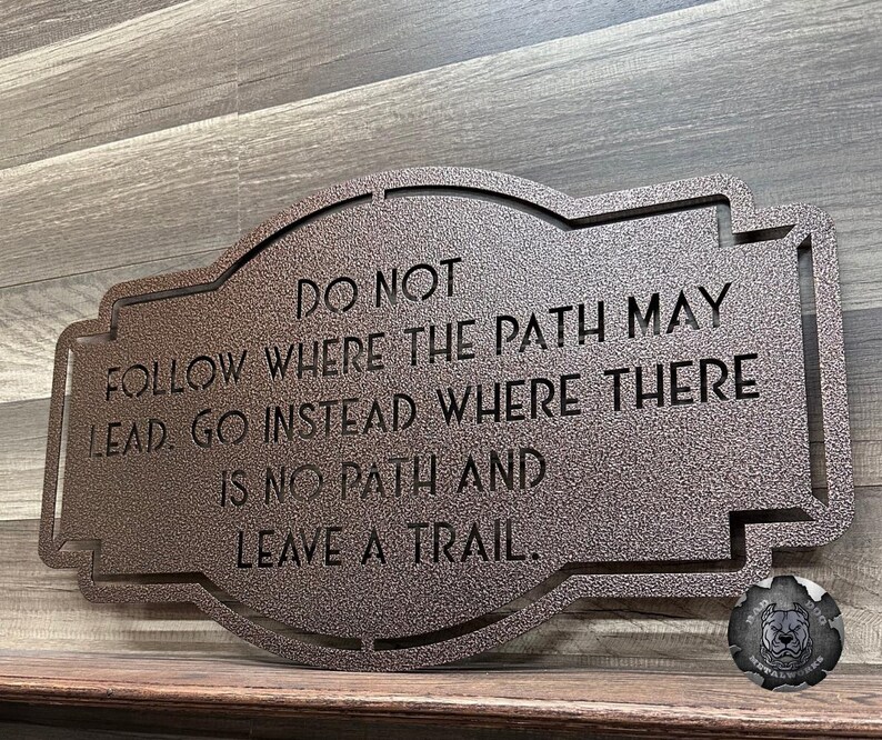Puede incluir: Un letrero de metal de color bronce con la cita: "NO SIGAS EL CAMINO. VE EN CAMBIO DONDE NO HAY CAMINO Y DEJA UN RASTRO." El letrero tiene un borde decorativo y es adecuado para exhibici&oacute;n en interiores o exteriores.