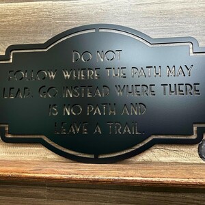 Puede incluir: Letrero de metal negro con la cita "NO SIGAS EL CAMINO, VE EN CAMBIO DONDE NO HAY CAMINO Y DEJA UN RASTRO". El letrero tiene un borde decorativo y se muestra sobre una superficie de madera.