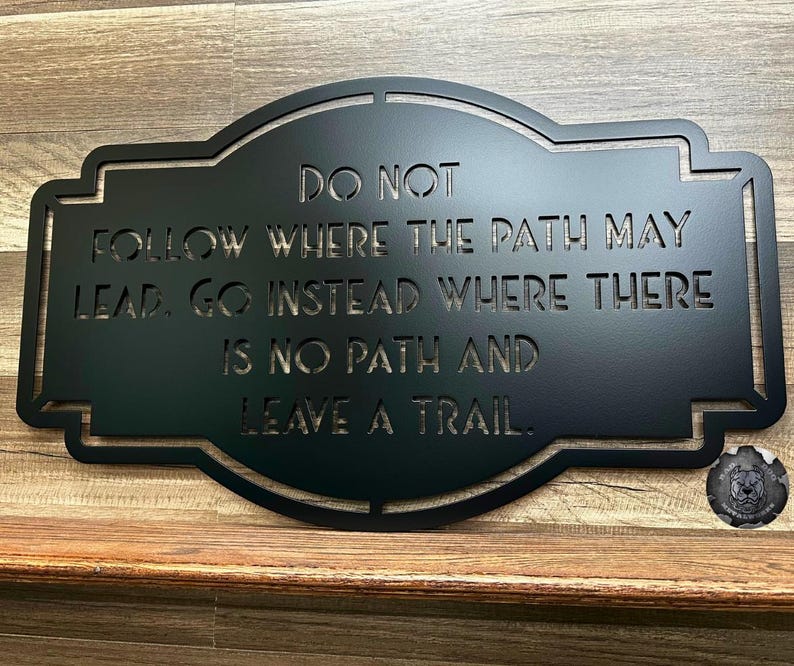 Puede incluir: Letrero de metal negro con la cita "NO SIGAS EL CAMINO, VE EN CAMBIO DONDE NO HAY CAMINO Y DEJA UN RASTRO". El letrero tiene un borde decorativo y se muestra sobre una superficie de madera.