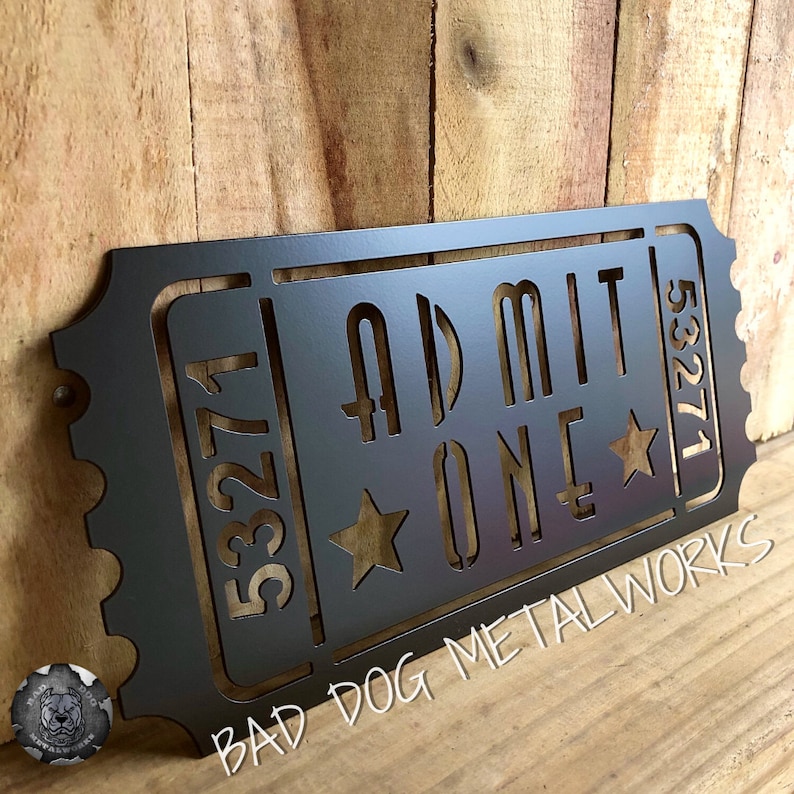May include: Black metal ticket-shaped sign with the text "ADMIT ONE" and the numbers "53271" and "53271" on either side. The sign also has two stars and the text "BAD DOG METALWORKS" at the bottom.