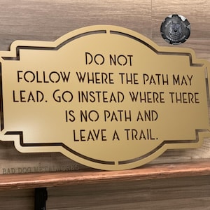 Puede incluir: Un letrero met&aacute;lico dorado con el texto "DO NOT FOLLOW WHERE THE PATH MAY LEAD. GO INSTEAD WHERE THERE IS NO PATH AND LEAVE A TRAIL." El letrero est&aacute; sobre una estanter&iacute;a de madera. Un emblema met&aacute;lico est&aacute; en la esquina superior derecha.