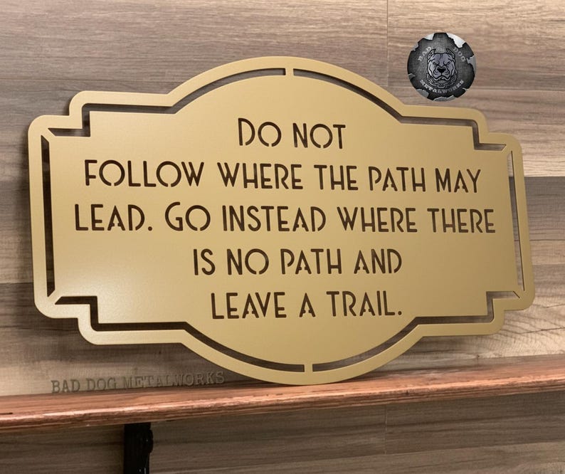 Puede incluir: Un letrero met&aacute;lico dorado con el texto "DO NOT FOLLOW WHERE THE PATH MAY LEAD. GO INSTEAD WHERE THERE IS NO PATH AND LEAVE A TRAIL." El letrero est&aacute; sobre una estanter&iacute;a de madera. Un emblema met&aacute;lico est&aacute; en la esquina superior derecha.