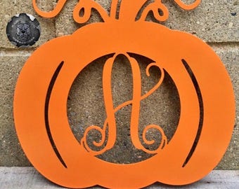May include: Orange metal pumpkin decoration with a cut-out letter "A" in the center. The pumpkin has a stem and curly vines. The decoration is likely for fall or Halloween.