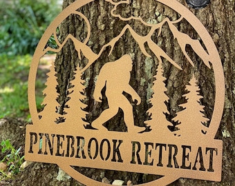 May include: A circular bronze-colored metal wall sign featuring a cutout silhouette of a Bigfoot figure walking through a forest of pine trees with mountains and clouds in the background. The text PINEBROOK RETREAT is embossed on a horizontal bar.