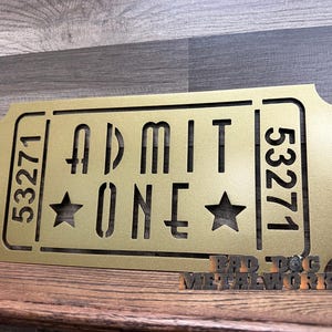 May include: Gold metal ticket-shaped sign with the text "ADMIT ONE" and the numbers "53271" on either side. Two stars are on either side of the word "ONE".
