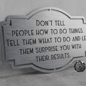 May include: A brushed silver metal wall sign with a decorative border featuring the cut-out text: DON'T TELL PEOPLE HOW TO DO THINGS. TELL THEM WHAT TO DO AND LET THEM SURPRISE YOU WITH THEIR RESULTS. A small circular logo is in the bottom right corner.