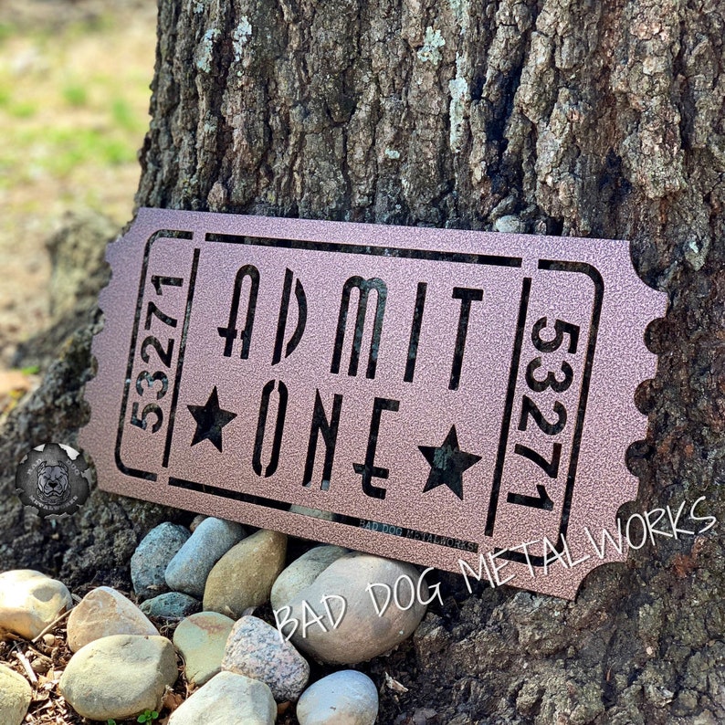 May include: A rusty brown metal sign shaped like a ticket stub with the text "ADMIT ONE" and the numbers "53271" on either side. The sign has two stars on either side of the word "ONE". The bottom of the sign reads "BAD DOG METAL WORKS".