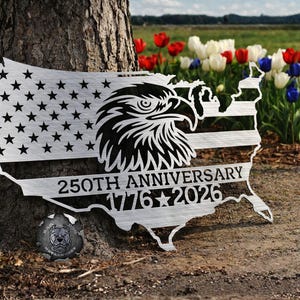May include: A metal wall art piece shaped like the United States, featuring the American flag design with stars and stripes. An eagle head is centered, and the text "250TH ANNIVERSARY 1776-2026" is displayed. The art is placed near a tree.