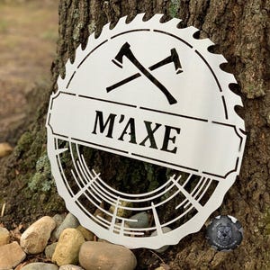 May include: A white metal sign shaped like a saw blade with two crossed axes and the text "M'AXE" in a banner.