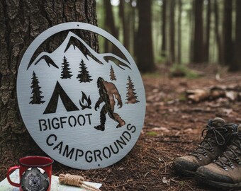 May include: A circular metal sign with a Bigfoot design, mountains, trees, and the words "BIGFOOT CAMPGROUNDS." A red enamel mug with a dog graphic and hiking boots are in the foreground, suggesting a camping theme.
