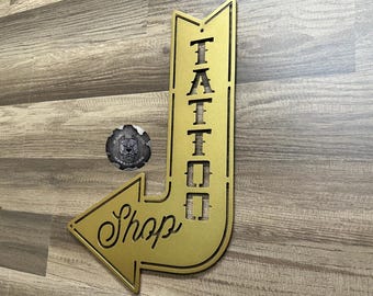 May include: Gold metal arrow sign with the word "Tattoo" vertically written on the left side and the word "Shop" written horizontally on the right side.