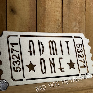 May include: A silver metal ticket with the words "ADMIT ONE" and the number "53271" on it. Two stars are on either side of the word "ONE". The ticket is on a wooden background. The words "BAD DOG METALWORKS" are at the bottom of the image.