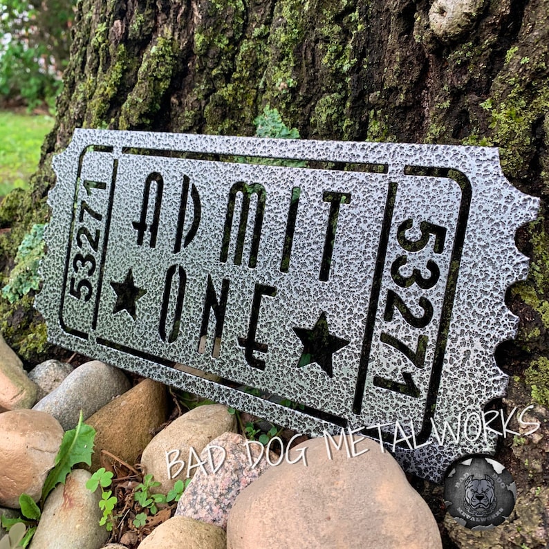 May include: A silver metal ticket with the text "ADMIT ONE" and the number "53271" on it. The ticket is textured and has two stars on it. The ticket is sitting on a bed of rocks.