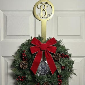 May include: A Christmas wreath with a red velvet bow, pinecones, and red berries. It hangs from a gold-colored metal door hanger with a decorative circular design and the letter "B". The wreath is displayed on a white door.