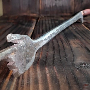 May include: Hand-forged metal tool with a unique shape, featuring a handle and a head with a fork-like design. The tool has a silver finish and a copper-colored end. The item is displayed on a dark wooden surface.