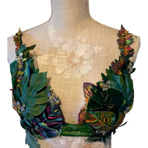 May include: A decorative bralette top adorned with layered artificial green tropical leaves, including monstera and fern shapes, accented with iridescent crystal embellishments and a dark fabric base featuring vibrant purple and pink streaks.