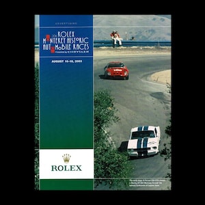 May include: Advertising poster for the 2003 Rolex Monterey Historic Automobile Races. The poster features a red Ferrari and a white and blue striped race car on a winding track. The Rolex logo is at the bottom.
