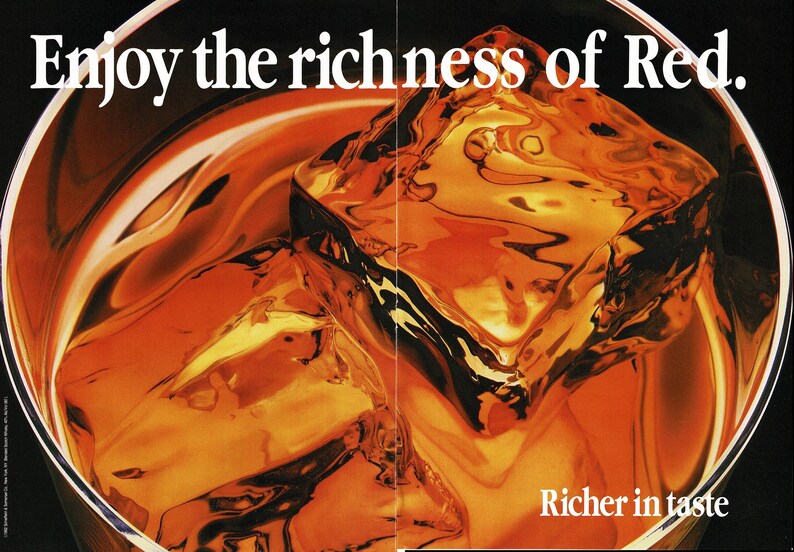 May include: Close-up of a glass filled with amber liquid and ice cubes. The text "Enjoy the richness of Red" is at the top, with "Richer in taste" at the bottom. The drink appears to be a whiskey or similar spirit.