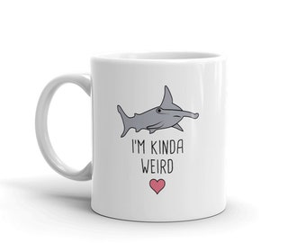 Può includere: Tazza in ceramica bianca con uno squalo martello disegnato e il testo "I'm kinda weird" con un cuore rosa.