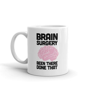 Puede incluir: Taza de café de cerámica blanca con un diseño en negro y rosa. La taza presenta el texto "Brain Surgery Been There Done That" y un gráfico de cerebro rosa.
