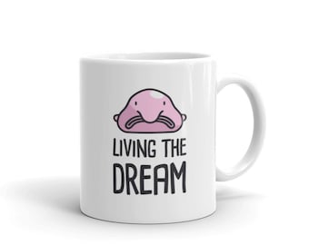 May include: White ceramic mug featuring a cartoon blobfish illustration and the text "Living the Dream".