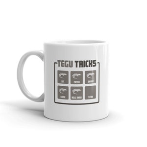 May include: White ceramic mug featuring a black and grey graphic of a Tegu lizard with the text "Tegu Tricks" and a grid of six commands: Sit, Fetch, Shake, Come, Roll Over, Stay.