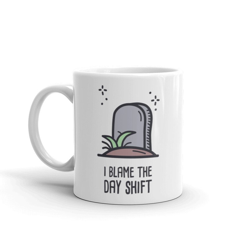 May include: White ceramic mug with a black handle featuring a cartoon illustration of a tombstone with grass and the text "I Blame The Day Shift". The design includes small starbursts.
