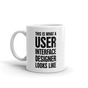 May include: White ceramic mug with a curved handle. The mug features the text "THIS IS WHAT A USER INTERFACE DESIGNER LOOKS LIKE" in bold black capital letters, making it a humorous gift for UI designers.