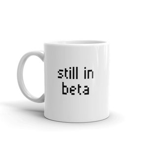 May include: White ceramic mug with black text that reads "still in beta".