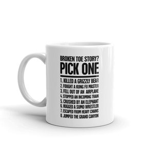May include: White ceramic mug with a black text that reads "BROKEN TOE STORY? PICK ONE" followed by a list of humorous options, including "Killed a Grizzly Bear" and "Jumped the Grand Canyon."