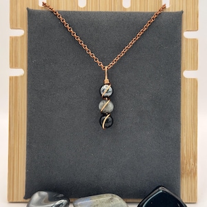 May include: A copper chain necklace with three gemstone beads: a black, a gray, and a black and gray bead. The beads are wire-wrapped in gold-colored wire. The necklace is displayed on a gray background with a wooden stand.