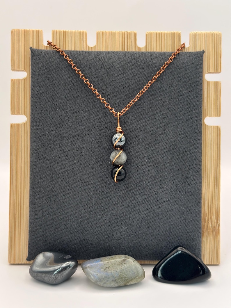 May include: A copper chain necklace with three gemstone beads: a black, a gray, and a black and gray bead. The beads are wire-wrapped in gold-colored wire. The necklace is displayed on a gray background with a wooden stand.